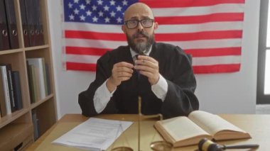 Amerikan bayrağı geçmişi olan, elinde kalem tutan ve tokmak ve hukuk kitaplarının yanında gözlük ve mahkeme cübbesi takan bir erkek hakim.