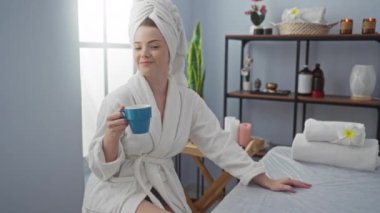 Bir kadın spa odasında rahatlatıcı bir anın tadını çıkarıyor, beyaz bir bornoz giyiyor ve elinde mavi bir kupa tutuyor, etrafı güzellik dekoruyla çevrili.