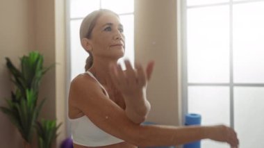 Olgun bir kadın spor salonunda sakin bir ifadeyle egzersiz yapıyor, gündüz antrenmanı sırasında spor aletleri ve bitkilerle çevrili yoga hareketleri yapıyor..