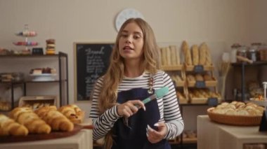 Sıcak bir pastanede içi ekmek ve hamur işleriyle dolu, çizgili üst ve mavi önlüklü sıcak ve davetkar bir atmosferi canlandıran, taze kruvasan servis eden bir kadın..