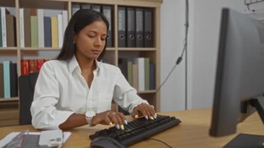 Ofiste çalışan, klavyede yazan, beyaz gömlek giyen, esmer, odaklı yüz ifadesi olan, modern çalışma ortamında dokümanlar ve klasörlerle çevrili bir kadın.