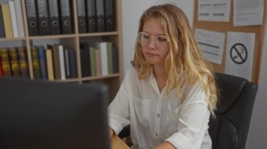 Ofis ortamında beyaz bir kadın bilgisayar ekranına bakıyor, kitaplarla ve ciltlerle çevrili, beyaz bir gömlek giyiyor, gözlüklü ve uzun sarı saçlı..