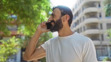 Güneşli şehir parkında telefonda konuşurken sakallı genç adam gülümsüyor. Arka planda belediye binaları var.