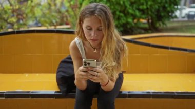 Şehir parkında sarı bir bankta oturan genç bir kadın akıllı telefonunda odaklanmış bir ifadeyle kayıyor.