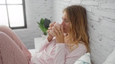 Sarı saçlı, rahat yatak odasında elinde fincanla oturan pembe pijama giyen ve pencerenin yanındaki beyaz tuğla duvara yaslanan genç bir kadın.