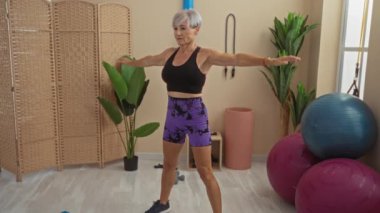 Yetişkin bir kadın spor salonunda spor yapıyor. Arka planda spor topları ve bitkiler var. Siyah bir bluz ve mor şort giyiyor.