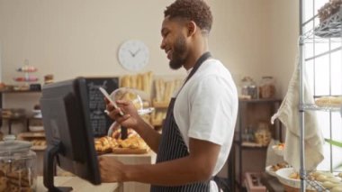Fırında çalışan genç adam akıllı telefon kullanırken gülümsüyor ve tezgahın arkasında raflar dolusu hamur işi ve ekmek ile duruyor.