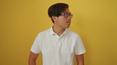 Gözlüklü, sarı arka planda duran, beyaz polo tişörtlü, çekici ve kendinden emin bir tavır sergileyen genç bir adam.