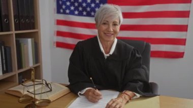 Ofisteki bir masada yazı yazan, kısa gri saçlı olgun bir kadın yargıç. Amerikan bayrağının arka planına karşı..