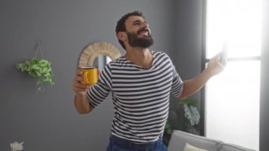 Sakallı genç adam evde gülüyor ve dans ediyor. Elinde sarı bir kupa ve akıllı bir telefonla modern dekor ve bitkilerle evde neşe ve mutluluk sergiliyor..