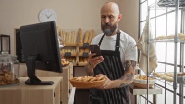 Sakallı İspanyol bir adam gün içinde fırında hamur işi fotoğrafı çekiyor, taze pişmiş ürünleri sergiliyor ve online içerik için akıllı telefon kullanıyor..