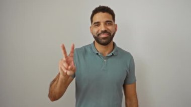 İzole edilmiş beyaz arka planda duran genç İspanyol adam, mutlu bir yüzle gülümsüyor, zafer işaretini jestle gösteriyor, galibiyeti iki parmakla kutluyor.