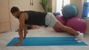 Spor salonunda denge topları ve bitkiler gibi spor aletleriyle dolu bir salonda egzersiz yaparken mavi bir paspasın üzerinde antrenman yapan bir adam.