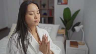 Bir kadın yatak odasında huzur içinde meditasyon yapıyor, beyaz bir bornoz giyiyor ve gözleri kapalı bir yatakta oturuyor, modern bir evin içinde sakin ve sakin bir atmosferi vurguluyor.