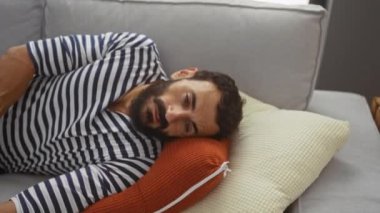 Sakallı genç adam, oturma odasındaki yastıklarla kanepede dinleniyor.