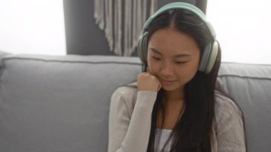 Oturma odasındaki koltukta oturan, kulaklık takan genç bir kadın, düşünceli bir ifadeyle.