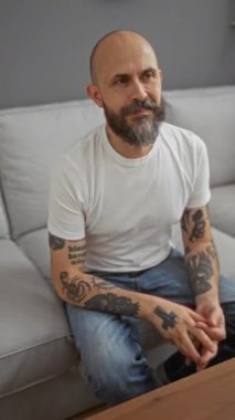 Oturma odasındaki gri bir koltukta oturan, kollarında dövmeleri olan, beyaz tişört ve kot pantolon giyen, kameraya doğru gülümseyen, iç mekan ayarlı bir adam.