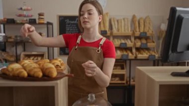 Kırmızı gömlek ve kahverengi önlükle arka planda ekmek ve hamur işleriyle kruvasan düzenleyen bir kadın.