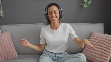 Genç bir kadın, rahat bir apartman dairesinde koltukta neşeyle dans ederken kulaklıkla müzik dinlemekten zevk alıyor.