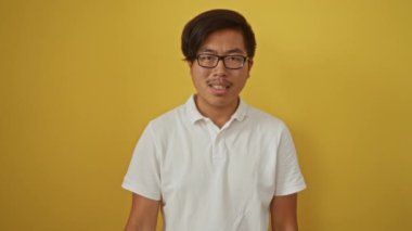 Gözlüklü beyaz gömlekli genç adam, izole edilmiş sarı arkaplanı onaylamıyor, ciddi bir ifadeyle memnuniyetsizlik veya hoşnutsuzluğu iletiyor..