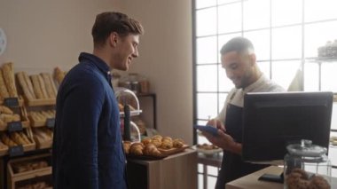 Bir pastanede çalışan adamlar, tabletten tablete bir şey gösteriyorlar, etrafları ekmek ve hamur işleriyle çevrili, kapalı bir dükkânda.