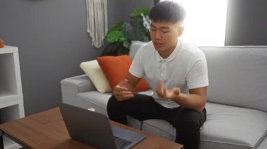 Video görüşmesi sırasında elinde hap şişesi olan genç adam, modern oturma odası ortamında gelişigüzel ve endişeli bir atmosferi vurguluyor, sağlık ve teknoloji etkileşimini vurguluyor..