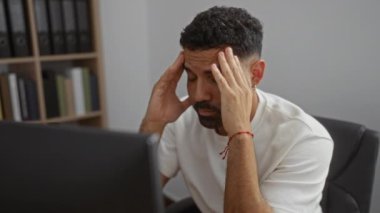 Ofisteki bir bilgisayara sinirle bakan genç bir adam, stres ve yorgunluk gösteren klasörlerin arkasında oturuyor.