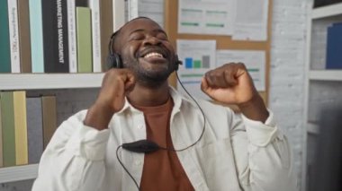 Kulaklık takmış, etrafı kitap ve belgelerle çevrili genç bir adam ofiste seviniyor.