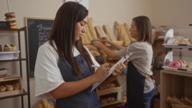 Pastanedeki kadın işçiler ekmek ve hamur işleri envanterini alırken, bir kadın gülümseyip pano tutarken diğeri malları düzenliyor.