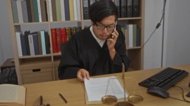 Siyah cüppeli genç adam, arka planda pulları ve kitaplığı olan yasal belgeleri okurken telefon kullanıyor..
