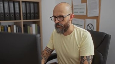 Ofis odasında çalışan İspanyol bir adam, ciddi bir ifadeyle bilgisayar görevlerine odaklanıyor, dosya ve kitaplarla dolu raflarla çevrili.