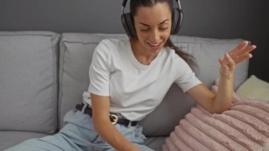 Kanepede dinlenen, kulaklık takan ve kot pantolon giyen, yanında yumuşak pembe yastıklı akıllı telefon kullanan bir kadın.