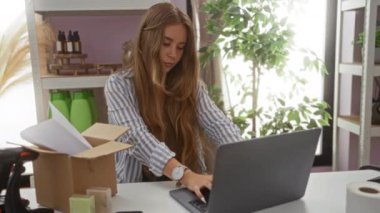 Ev dekorasyon mağazasında başarıyı kutlayan kadın, dizüstü bilgisayarda çalışıyor, masasında paketleme malzemeleri, bitkiler ve raflarla çevrili.