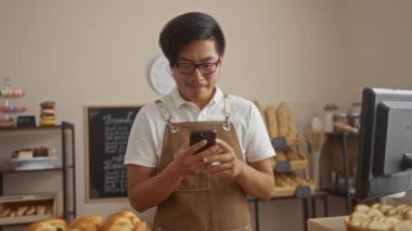 Pastanedeki genç Asyalı adam cep telefonunu kontrol ediyor. Fırında ekmek rafı yaparken heyecanlanıyor.