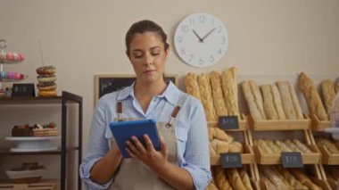 Fırında saat altında tablet kullanan çeşitli ekmeklerle genç bir kadın.