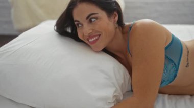 Mavi iç çamaşırlı, esmer, genç bir kadın rahat bir yatak odasında gülümsüyor ve yatakta uzanıyor.