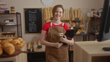 Ekmek, hamur işleri ve arka planda menü tahtasıyla dekore edilmiş fırında elinde pano tutan kadın fırıncı gülümsüyor..