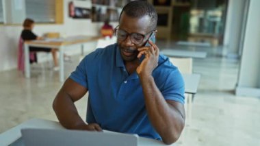 Ofiste, iş yerinde dizüstü bilgisayarda çalışırken telefonla konuşan genç adam, masa ve sandalyelerle iç mekanda ciddi ve profesyonel bir tavır sergiliyor..