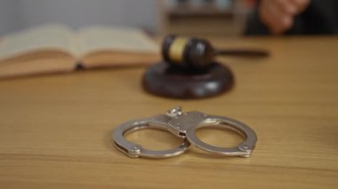 Tahta bir masaya kelepçelenmiş, açık bir hukuk kitabı ve mahkeme salonunda yargıç tokmağı var.