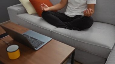 Evdeki kanepede dizüstü bilgisayarla meditasyon yapan bir adam modern oturma odasında huzurlu bir anı yakalıyor..