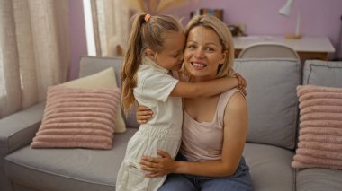 Anne ve kızı sıcak bir oturma odasında kucaklaşıyorlar, aile sevgisini ve sıcaklığını ev dekorunda somutlaştırıyorlar.