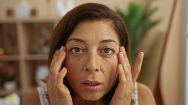 Orta yaşlı esmer bir kadın spa odasında tenini odaklanmış bir ifadeyle inceliyor. Güzelliği ve rahatlığı ön plana çıkarıyor..