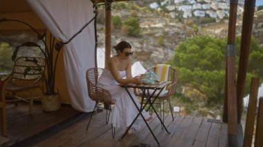 Güzel bir İspanyol genç esmer kadın Afrika 'nın dışında kamp yaparken görüldü. Etrafı doğa ile çevrili ahşap bir güvertede oturuyordu. Beyaz bir elbise giyiyordu ve akıllı telefonunu kullanıyordu..