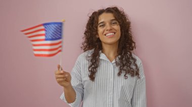 Amerikan bayrağı tutan genç bir kadın, gülümsüyor, pembe arka planda soyutlanmış, gururunu ve vatanseverliğini neşeli bir ifadeyle ifade ediyor.