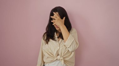 İzole edilmiş pembe arka planda genç bir kadın, elini yüzünü kapatıyor.