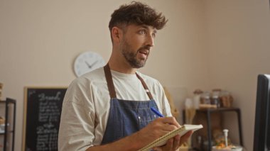 Fırında çalışan genç bir adam, arka planda raflar ve mallar olan kapalı bir dükkandaki not defterine notlar alıyor.