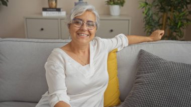 Oturma odasındaki rahat bir koltukta dinlenen gülümseyen bir kadın. Gri saçlı, olgun, beyaz, kapalı, iç mekan, yetişkin ve mutlu bir ortam..