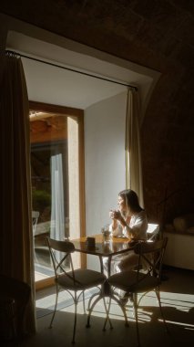Rahat bir otel ortamında huzurlu ve rahat bir atmosferi yansıtan, geniş pencerelerden içeri ışık saçan sakin bir kafeteryada kahveden zevk alan genç bir kadın..