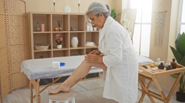 Gri saçlı yaşlı bir kadın, sağlık ürünleri ve dekor ile çevrili sakin bir spa odasında bacağına losyon sürüyor. Rahatlama ve kendine bakmanın önemini vurguluyor.