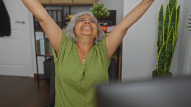 Ofiste kolları havada, yeşil gömlek ve gözlük takan, gri saçlı, yaşlı, gülümseyen, kapalı, mutlu, iş yerinde, olgun bir kadın kutluyor.